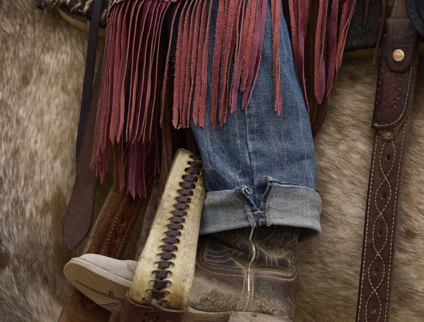 Zoomed in photo of a leg resting in a saddle with red chaps and jeans.
