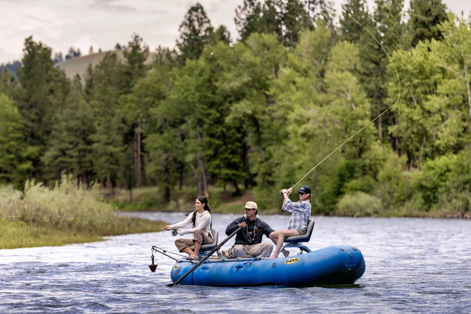 Two women fly fishing in a blue raft with a guide rowing.