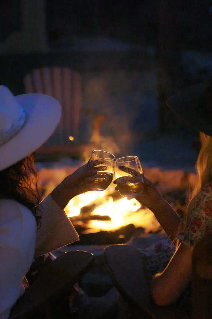 Two women clinking glasses of wine at night with a fire in the background.