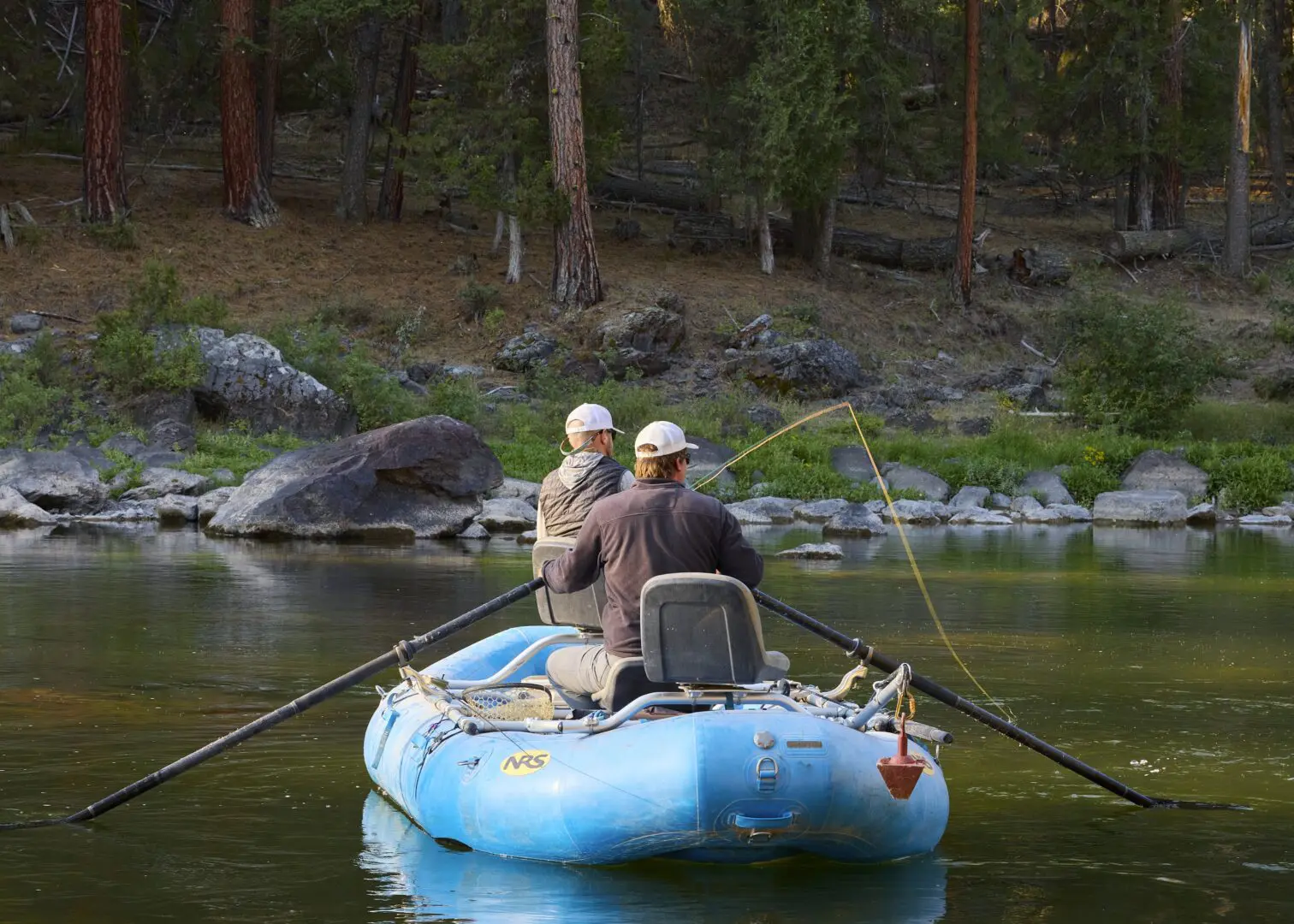 Two men fly fishing in a blue raft on a river.