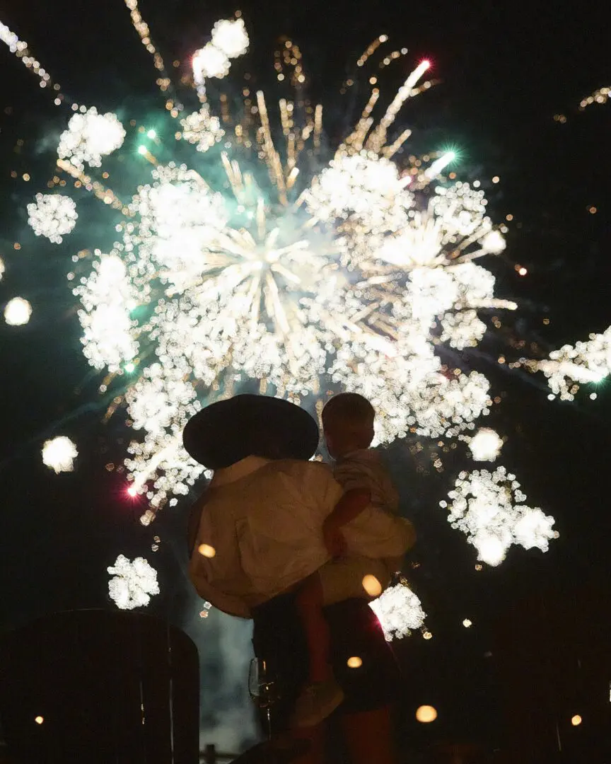 Woman holding a child with fireworks behind them.
