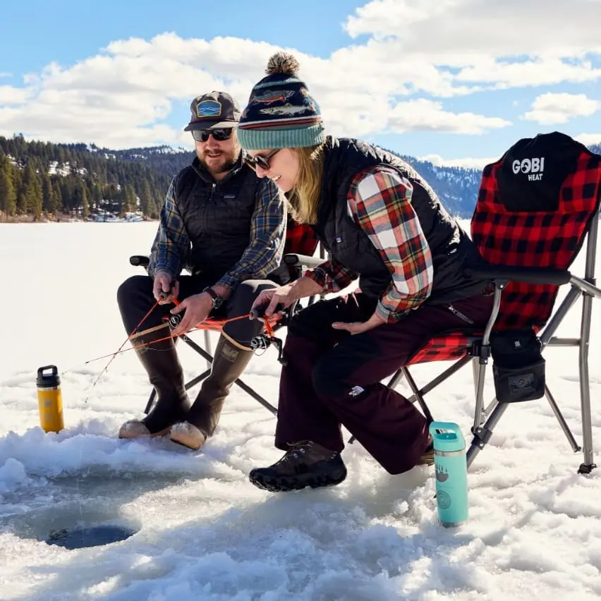 Two people in foldable chairs sitting on a frozen mountain lake, fishing into a small hole in the ice