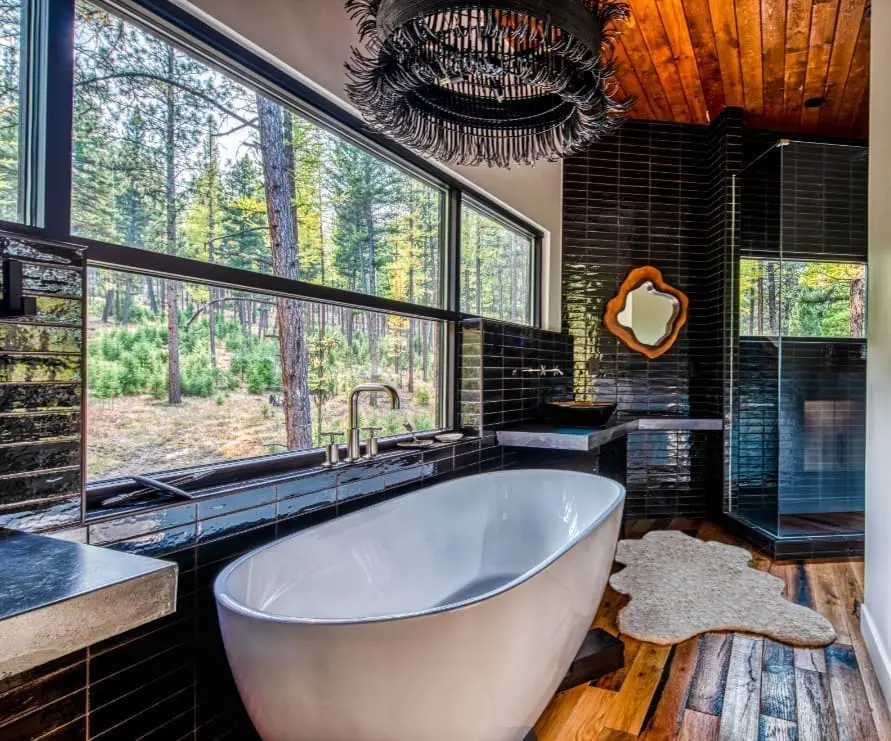Bathroom in the Tree Haus at Green O, featuring dark tiled wall, a bathtub under a modern chandelier, and a bright forest view from the window