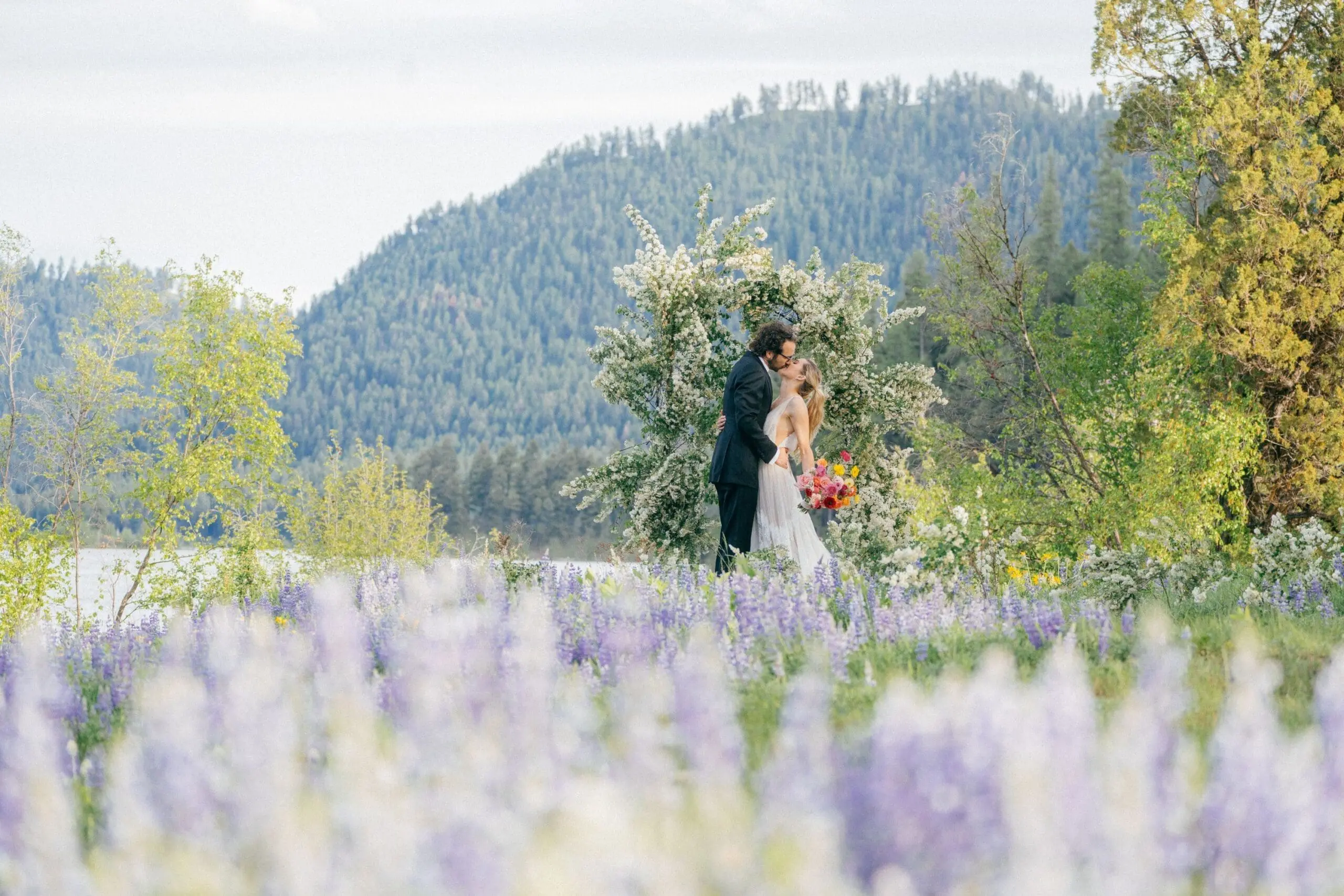 Bride and groom kissing under a flower arch on their spring wedding day surrounded by flowers and moutain greenery