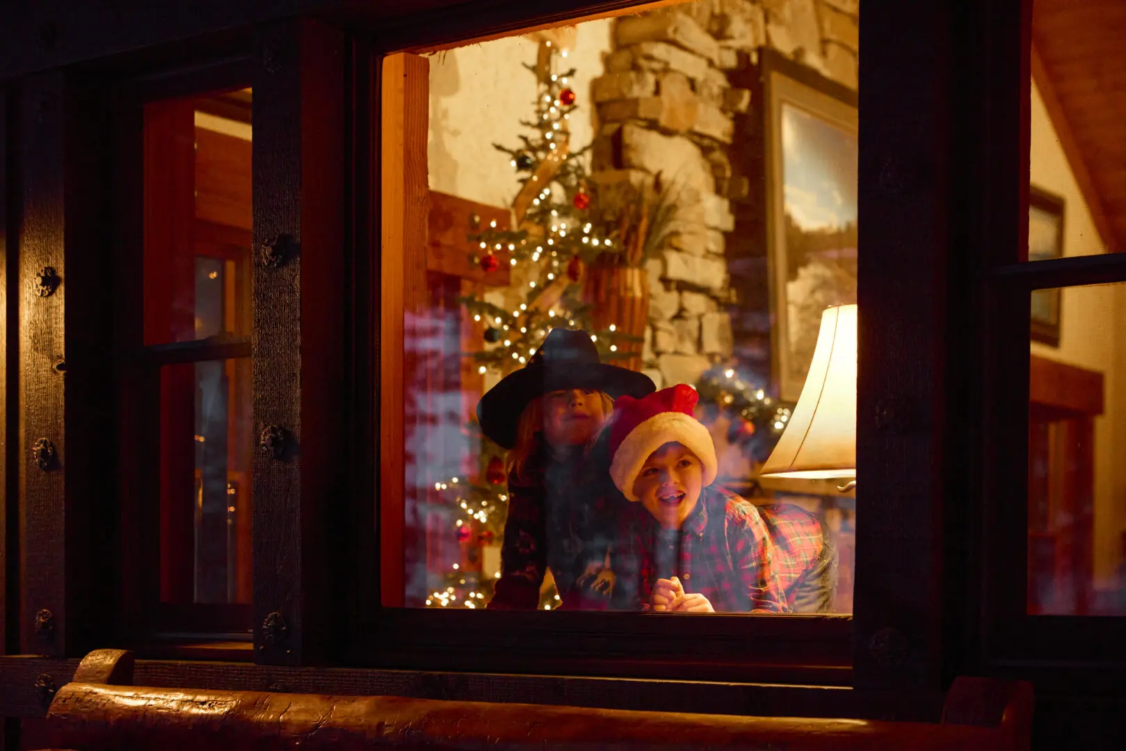 Two children looking out a window, one wearing a cowboy hat and the other a Santa hat.