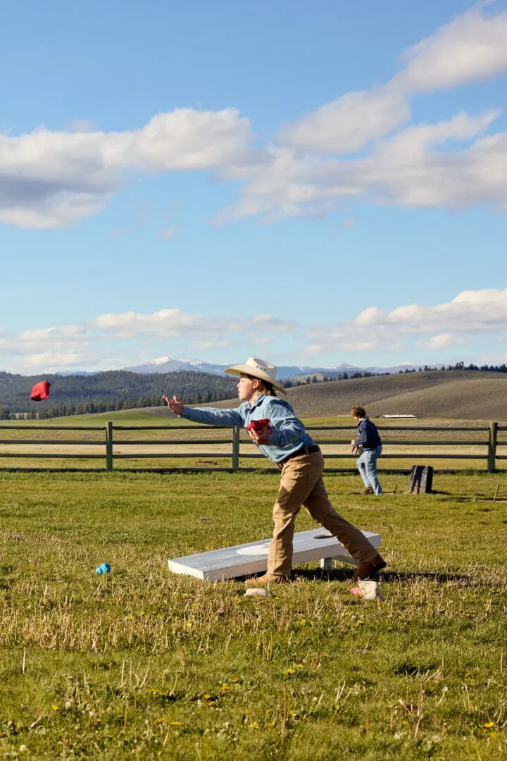 Boy in a cowboy hat playing cornhole outside.