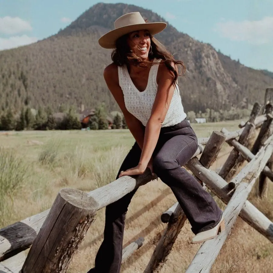 Woman sitting on a fence with a mountain in the background, wearing a hat, white tank top, and jeans.