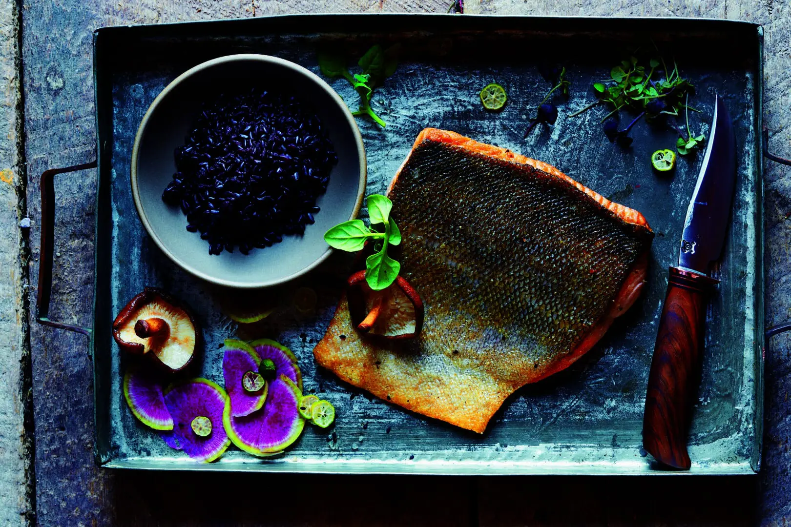 Cavier in a bowl, cooked fish, and radishes next to a knife, all laid out on a rectangular dinner platter.
