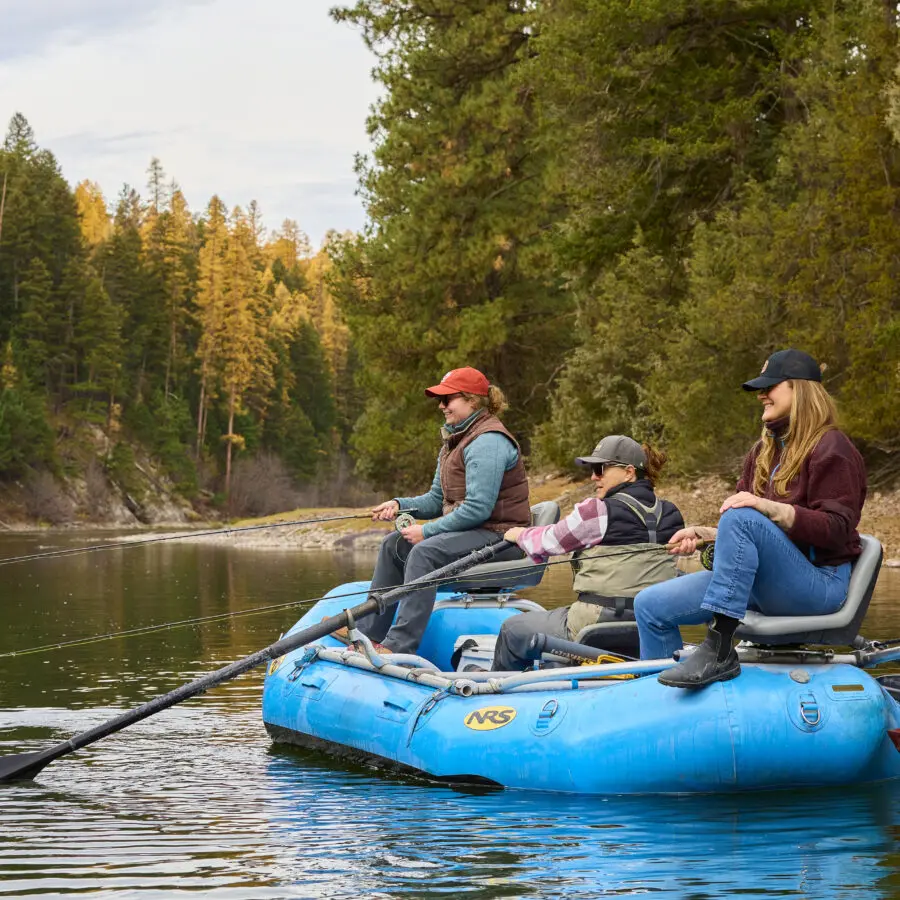 Three women fly fishing in a raft on a river with fall colored trees in the background.