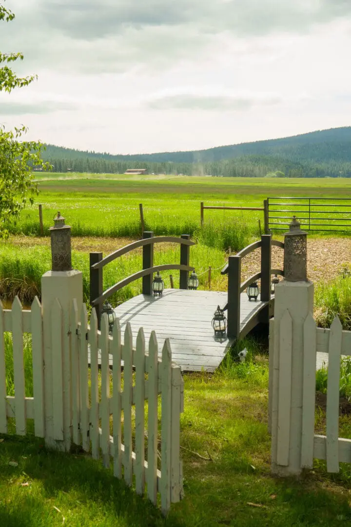 Green lawn with a white fence opening to a mini wooden bridge leading to the green yard.