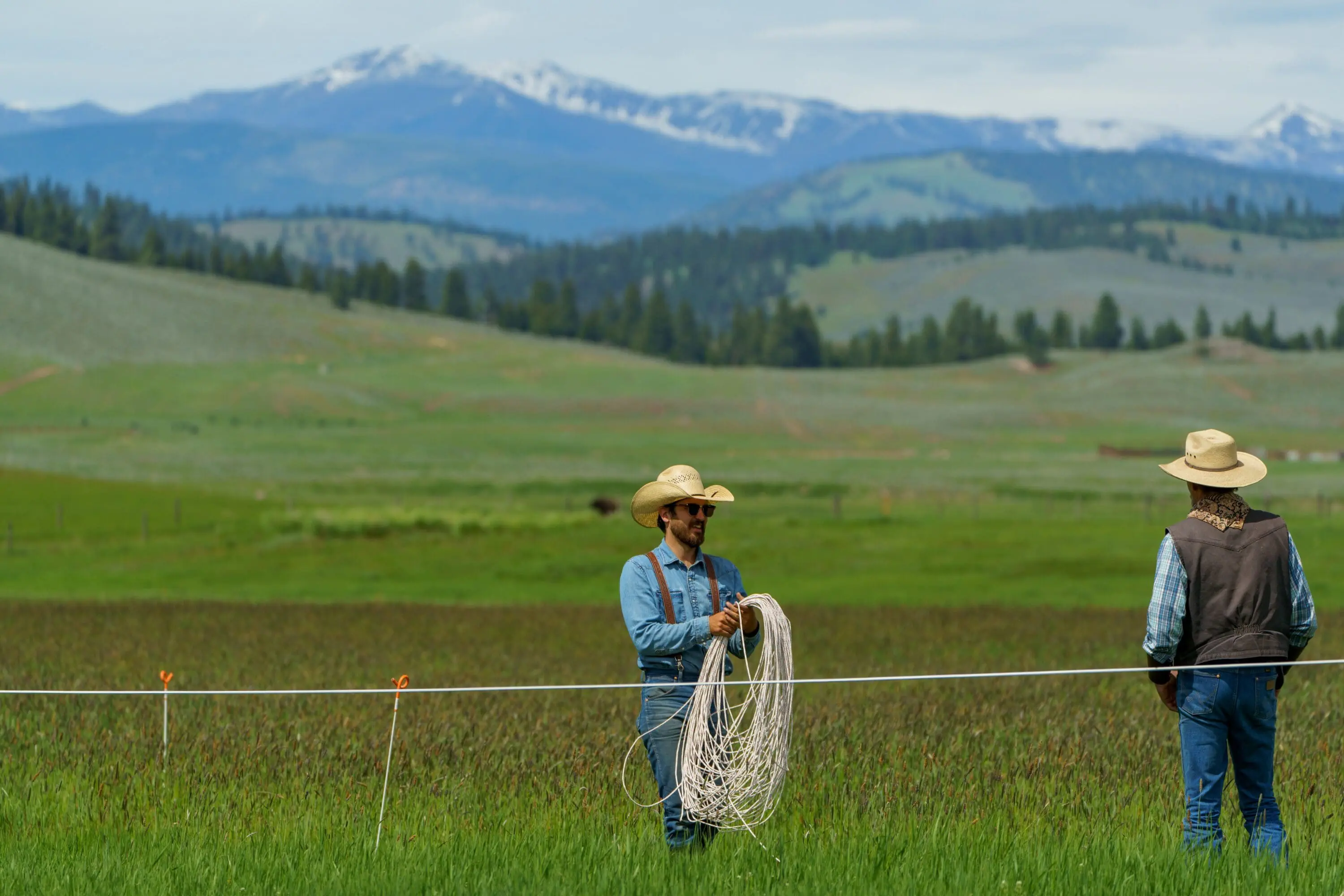 Two men adding wire fencing outside by a field and moutanins.