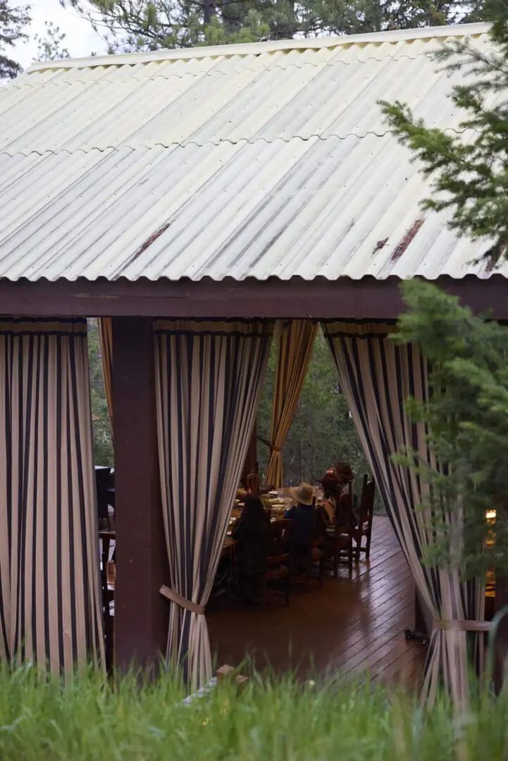 A pavilion with a tin roof and the curtains drawn back inside to show people sitting at a table indoors.