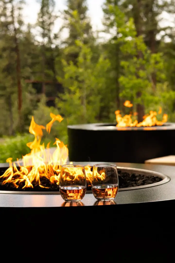 Two drinks sitting on the edge of a black fire pit with a live fire.