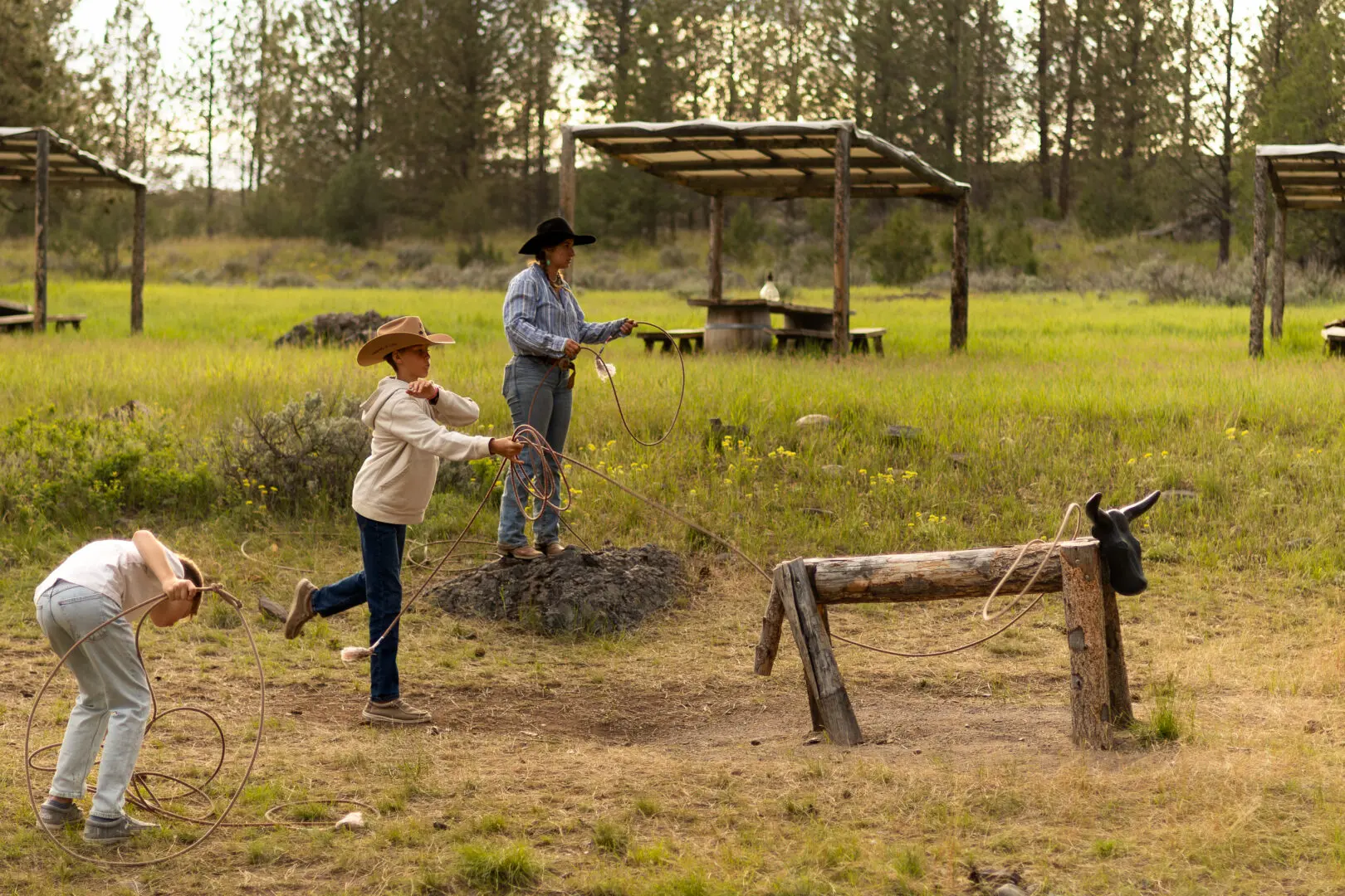 A child and parent playing western roping games on a fake wooden cattle outside in a grassy lawn.