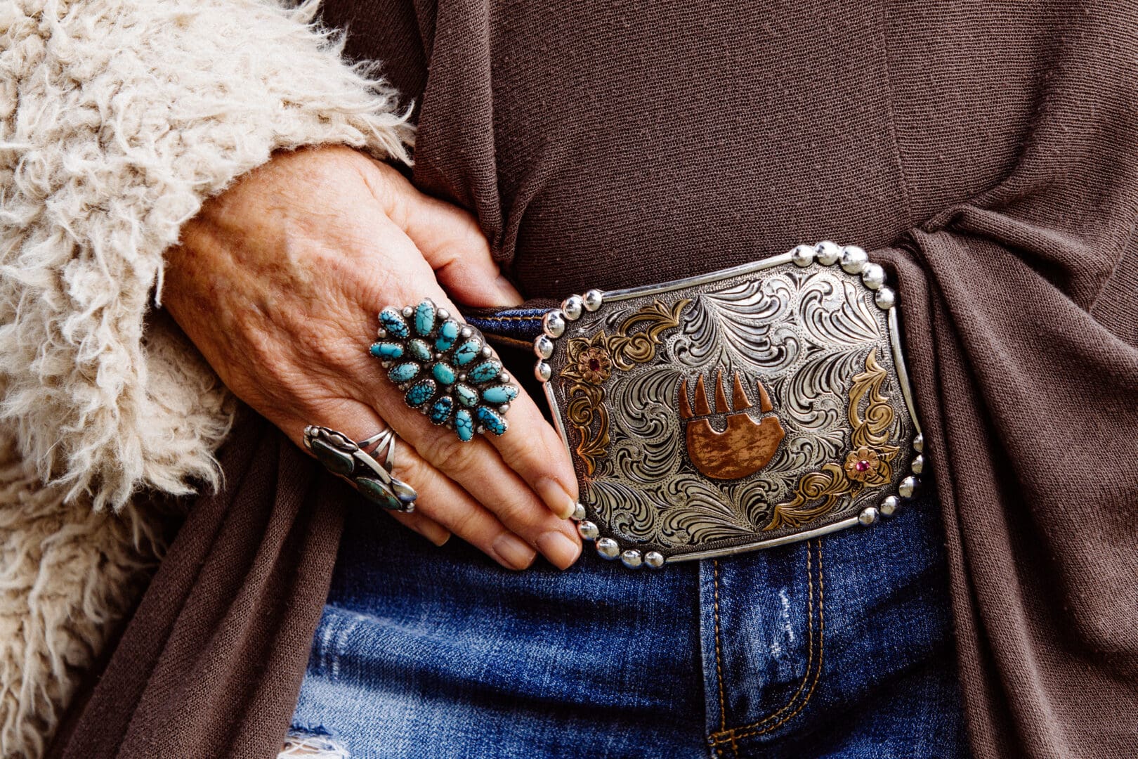 Womans hand resting on her western belt with a Paws Up Montana symbol, and turquoise rings.