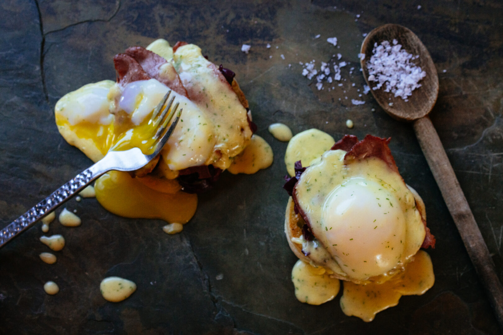 Two eggs Benedicts laid out on a table with salt and a fork piercing one of them.