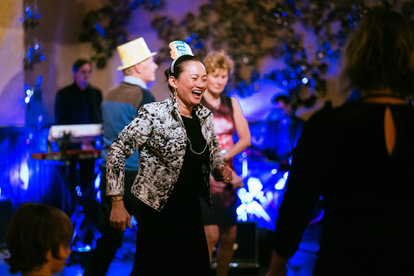 Woman dancing with new years eve hat on her head and other people dancing behind her.