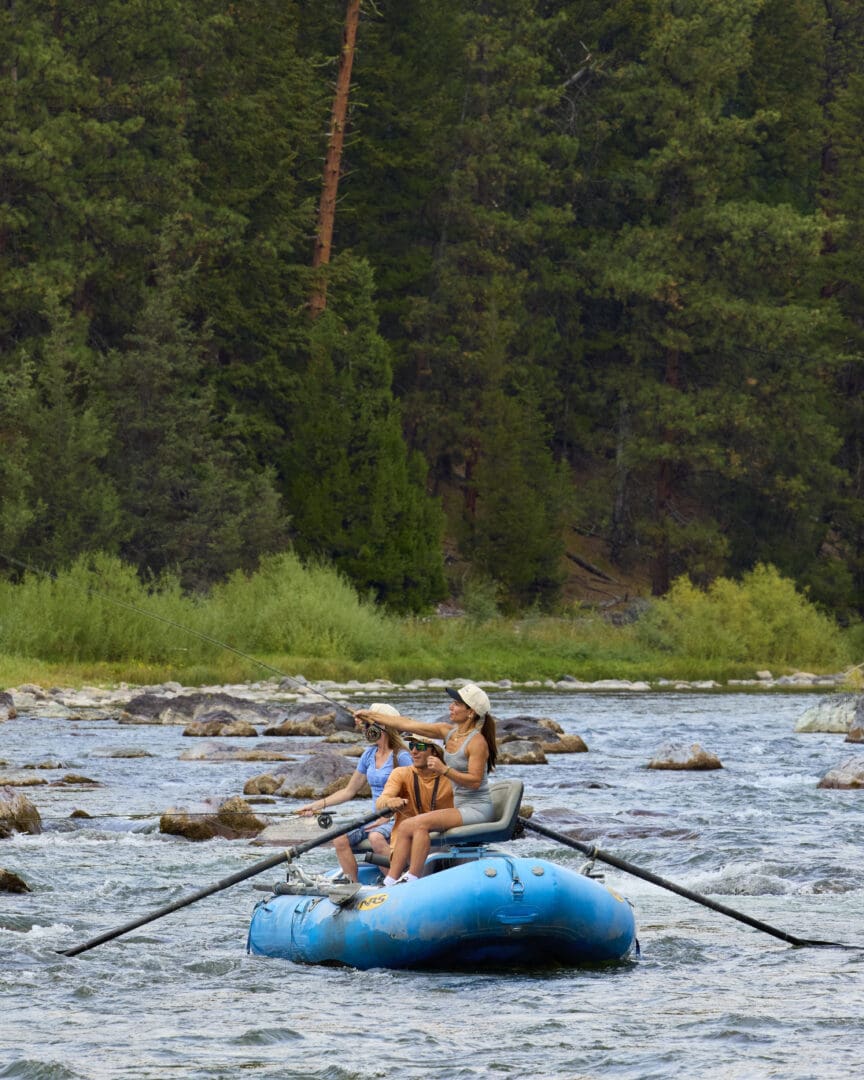 Women and man sitting in a raft floating down a river, while the woman fly fishes.