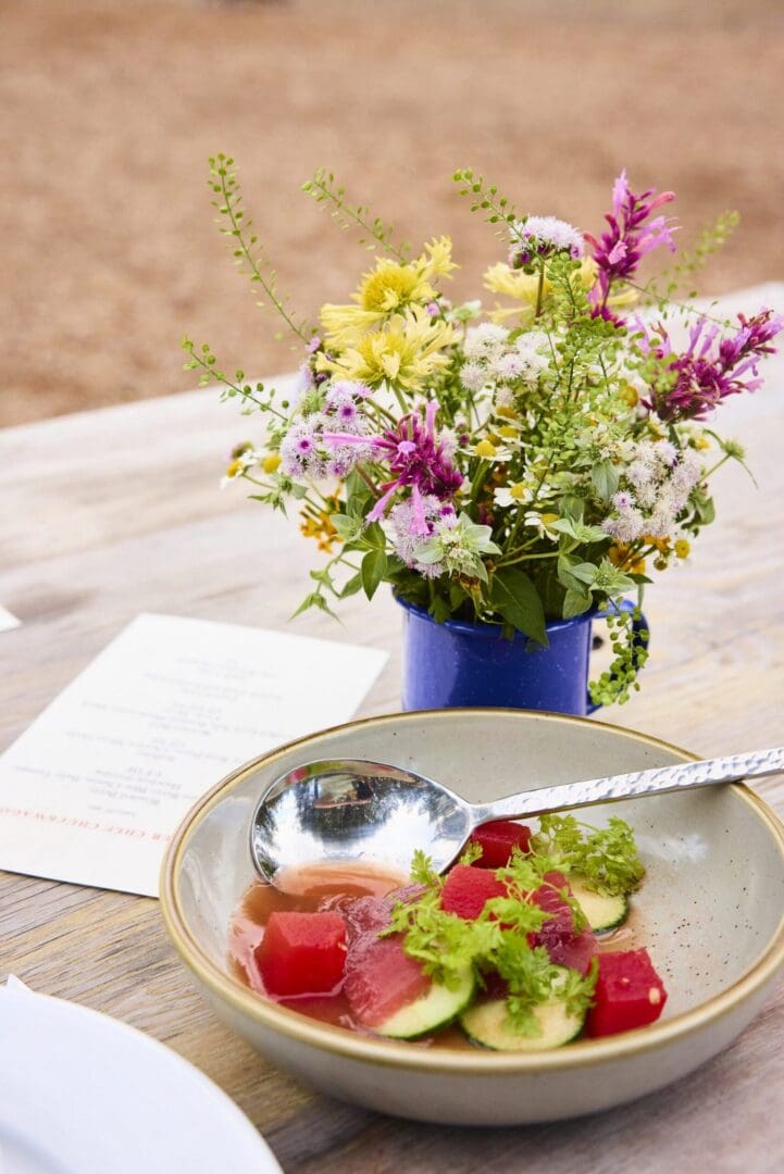 Watermelon fancy dish in a bowl with a spoon resting on a table with flowers beside it.