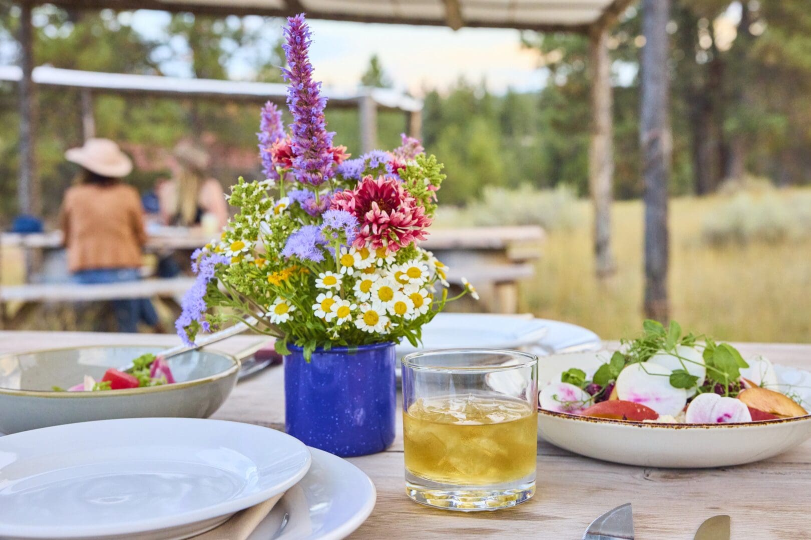 A picnic table outside set with flowers, plates, a salad, and a bright cocktail.