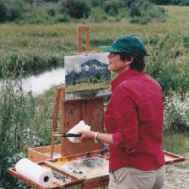 Woman in a redshirt paining outside behind mountains and a forest.