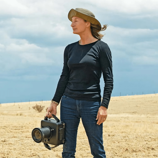 Woman holding a camera on a dry plain with a blue cloudy sky behind her.