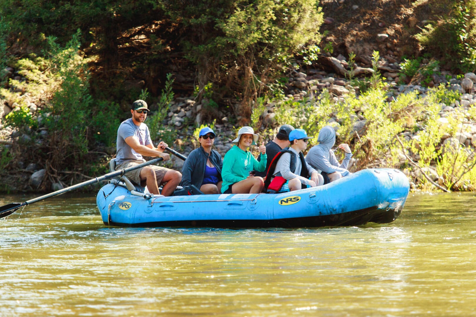 Group of people sitting in a blue raft while someone uses oars to push them down a river.