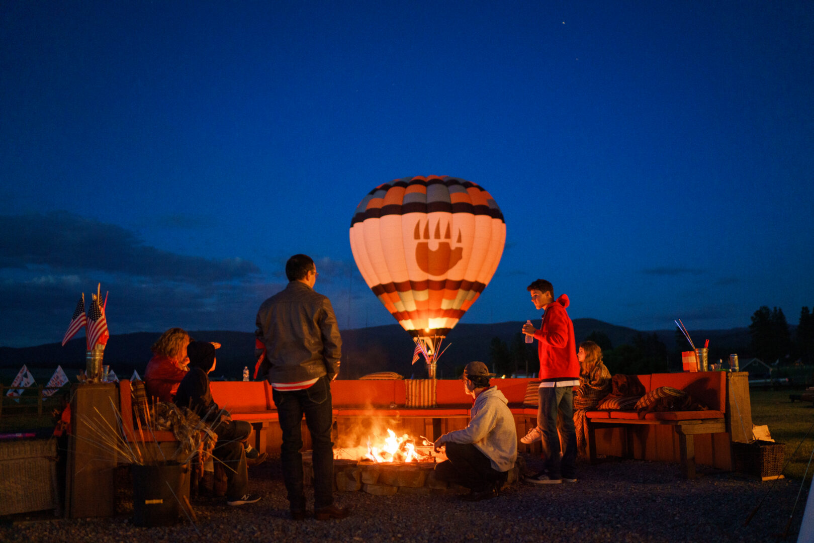 Family sitting around a fire with a hot air balloon in the background.