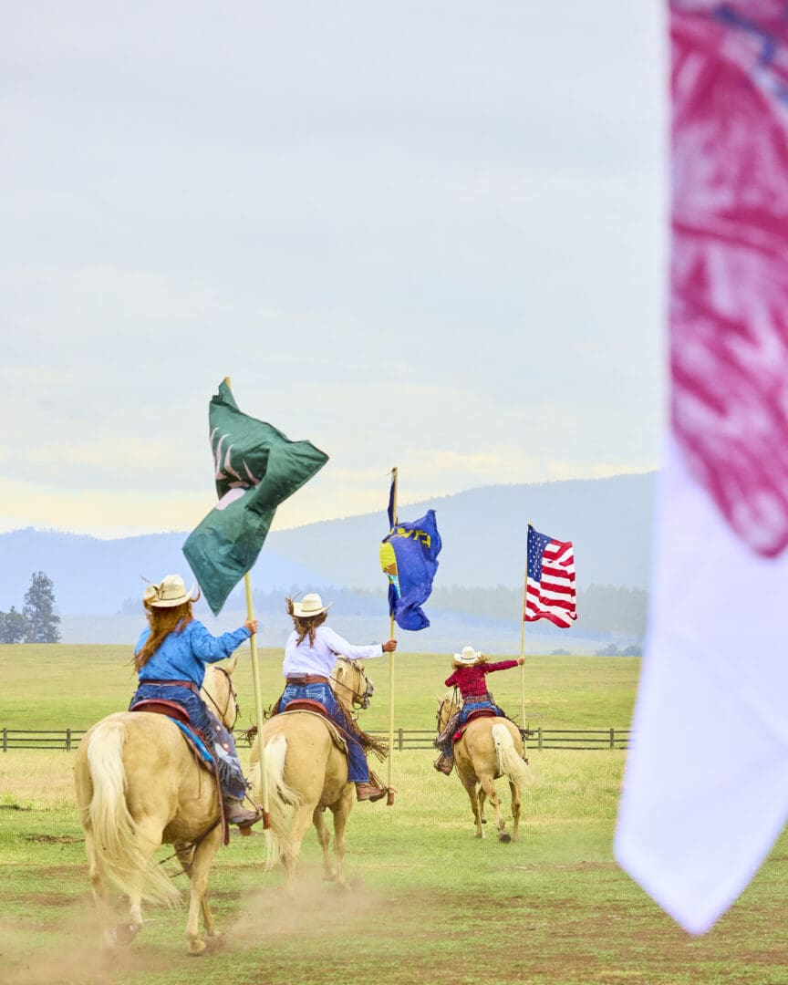 Three women on horses holding different flags with a green prairie and mountains in the background.