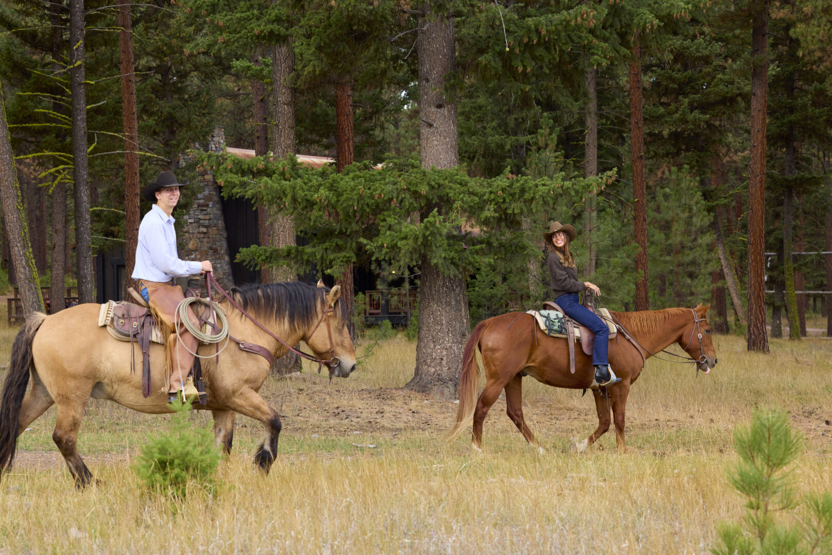 Man and woman riding horses with pines and trees in the background.