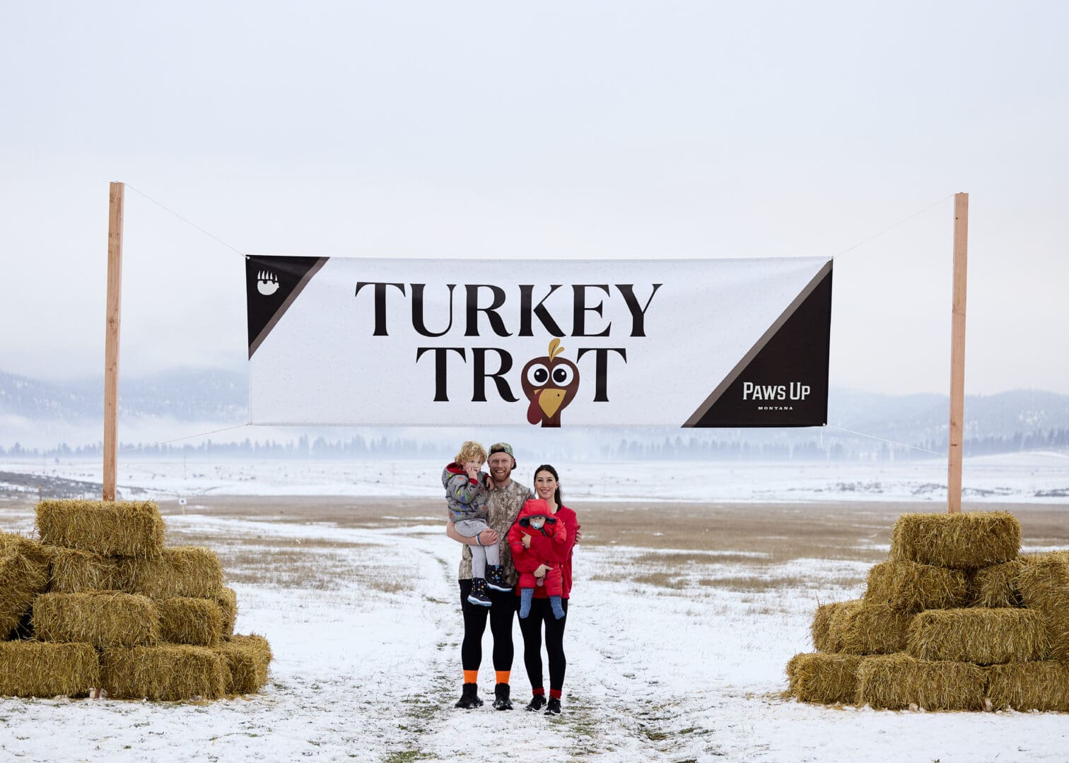 A family of three standing in front of a Turkey Trot sign with hay bails.