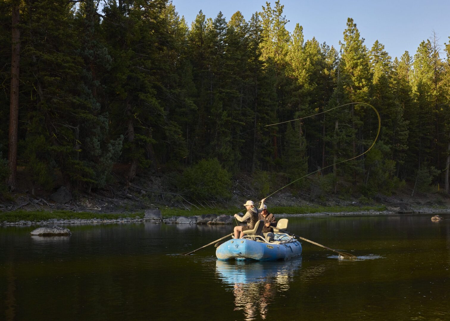 Men fly fishing on a river with trees behind them.
