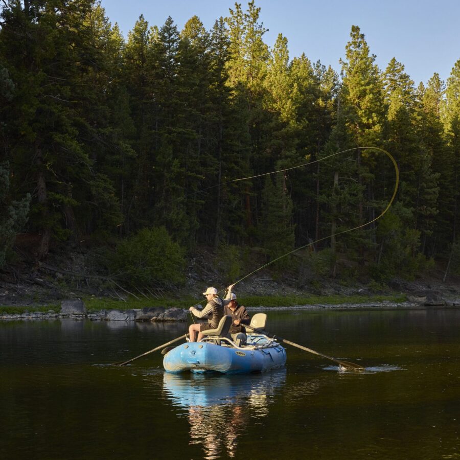 Men fly fishing on a river with trees behind them.