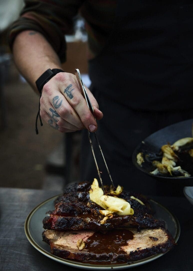 Chef preparing a meat dish with the tattooed word "chef" on his knuckles.