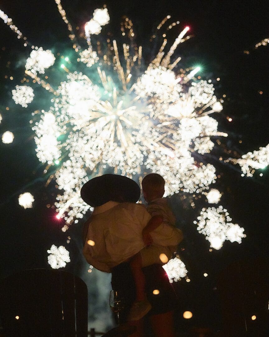 Woman holding a child with fireworks behind them.