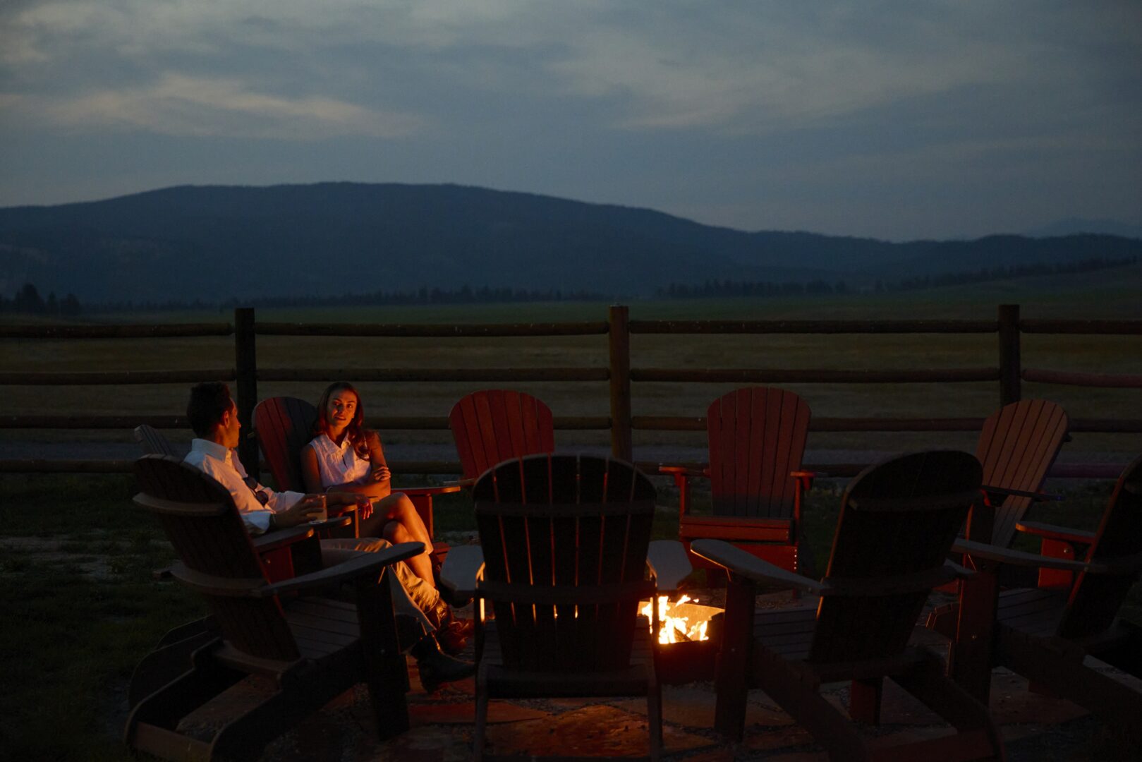 People sitting around a fire at dusk with mountains in the background.