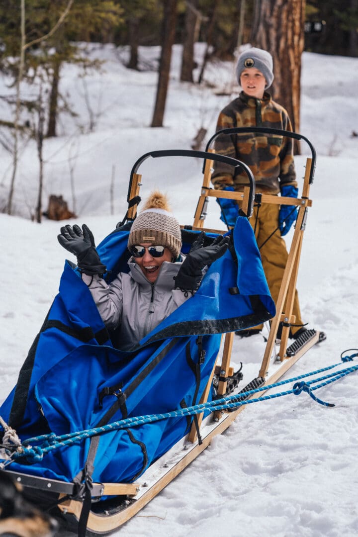 Boy sitting on the back of a dog sled with a woman riding in front under a blue sled cover
