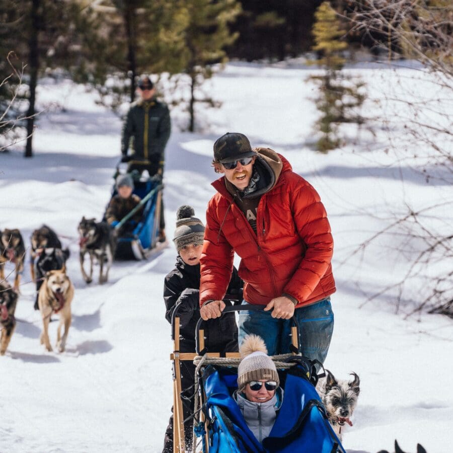 Man and child riding on the back of a dog sled with a woman sitting in front riding it on a snowy day.