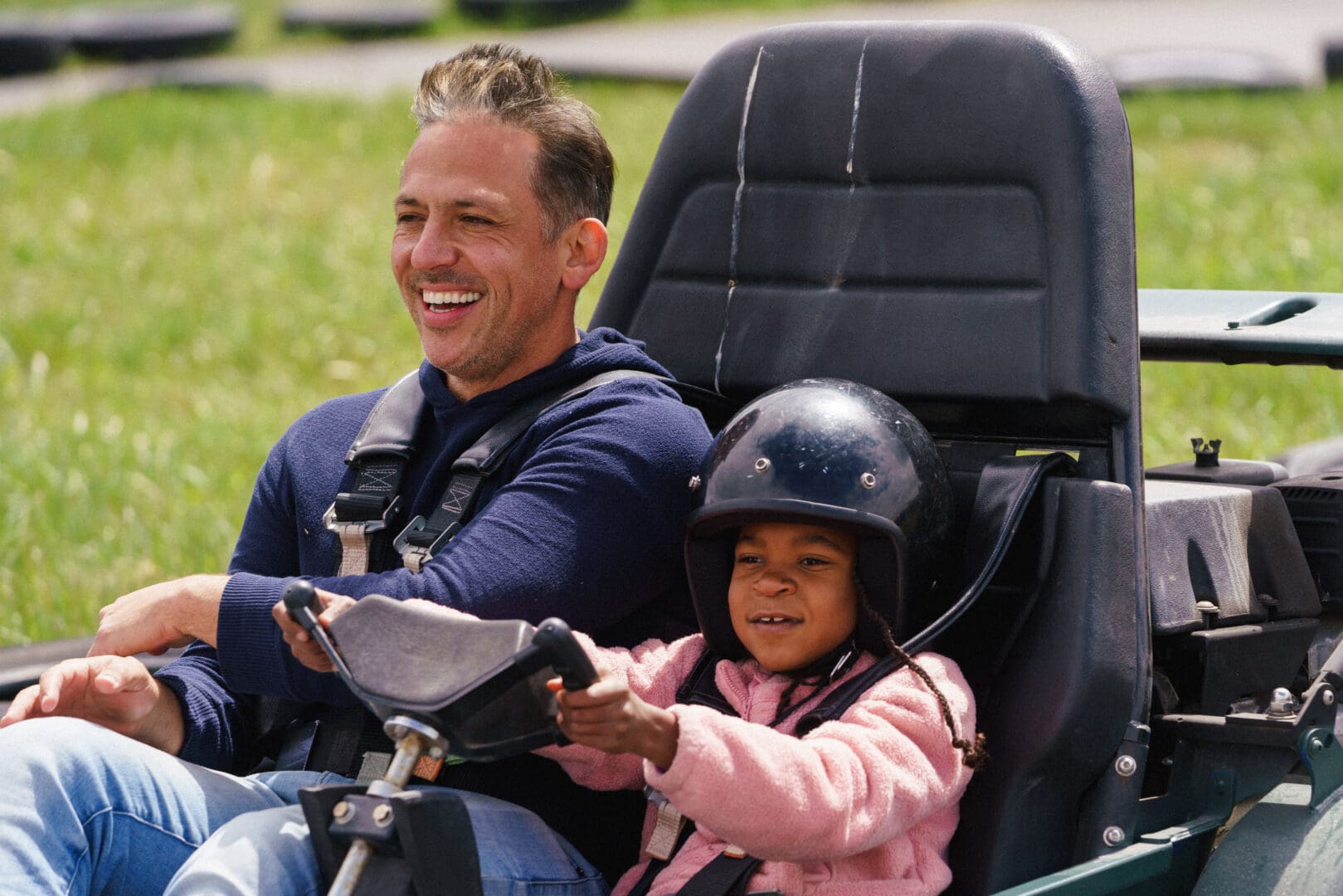 Father and daughter sitting in a go-kart together getting ready to race on an outdoor track.
