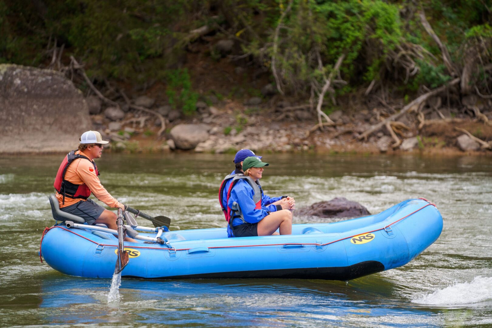 Two people sitting in a blue raft while a guide uses oars to push them down a river.