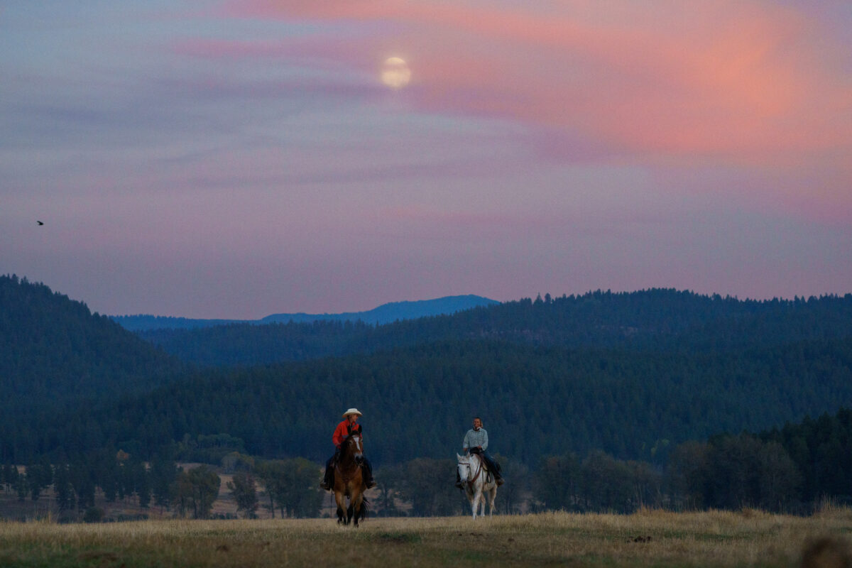 Two riders riding horses at sunset with the moon in the background and mountains.