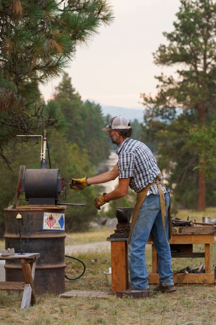 Man working at a iron stove outside surrounded by trees.