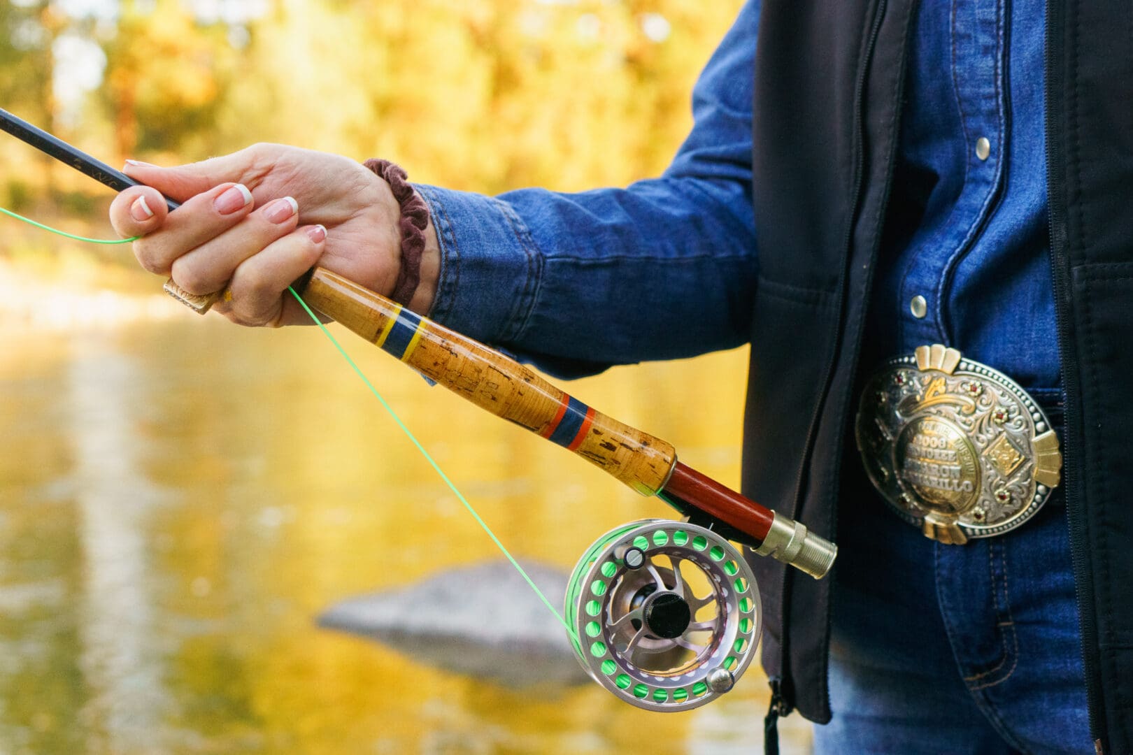 Man wearing a western belt holding a fishing rod by a river.