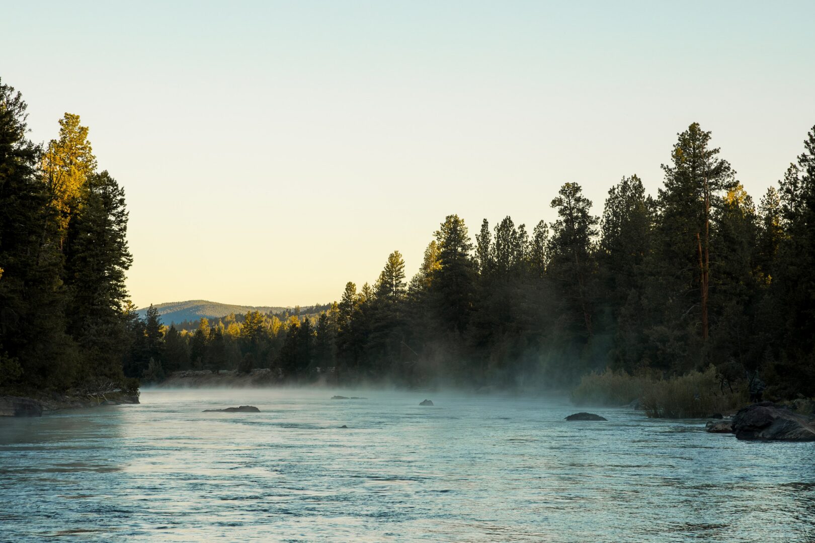A running river with steam rising off of it with trees on either side.