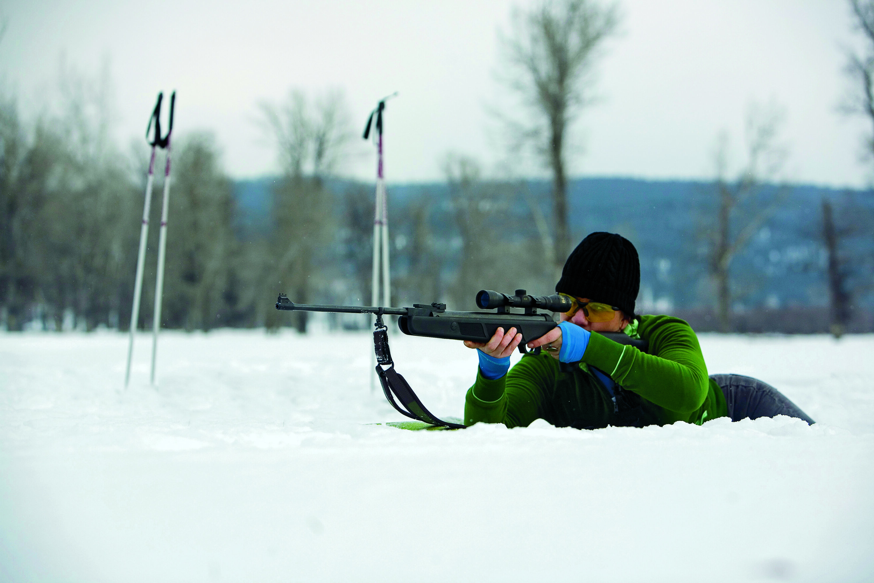 Woman in a green winter outfit laying in the snow to shoot a gun.