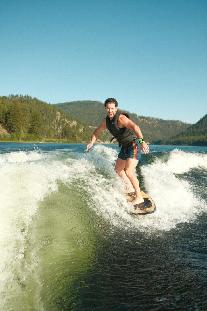 Man on a wake board in the middle of a lake with trees and hills in the background.