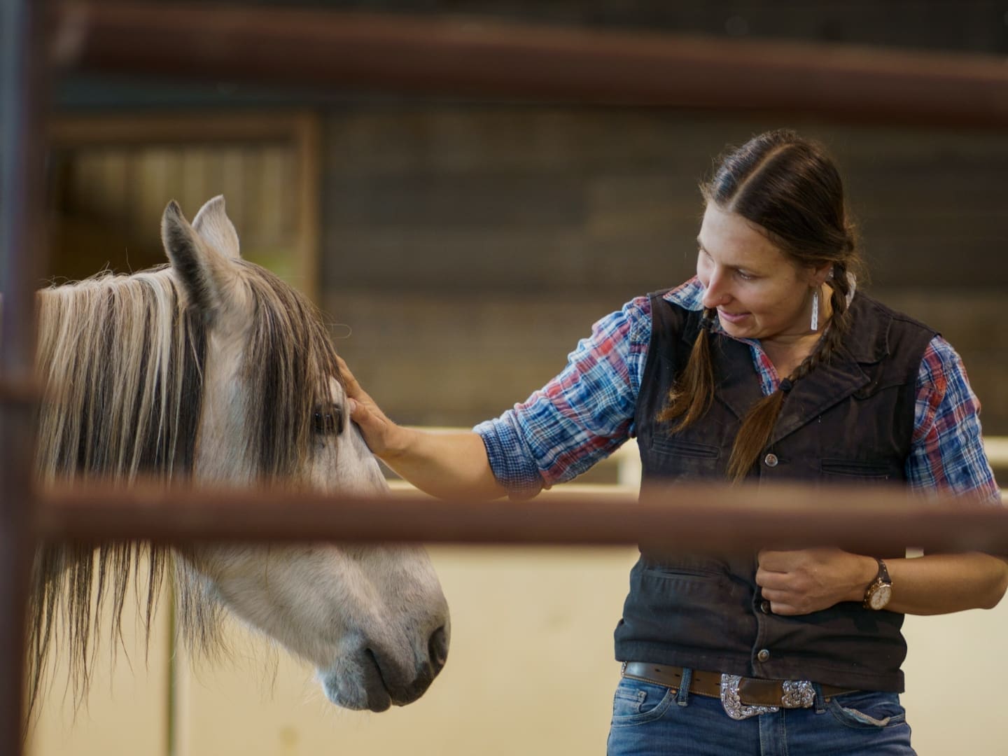 Cowgirl talking to and petting a white and gray horse in an equestrian arena