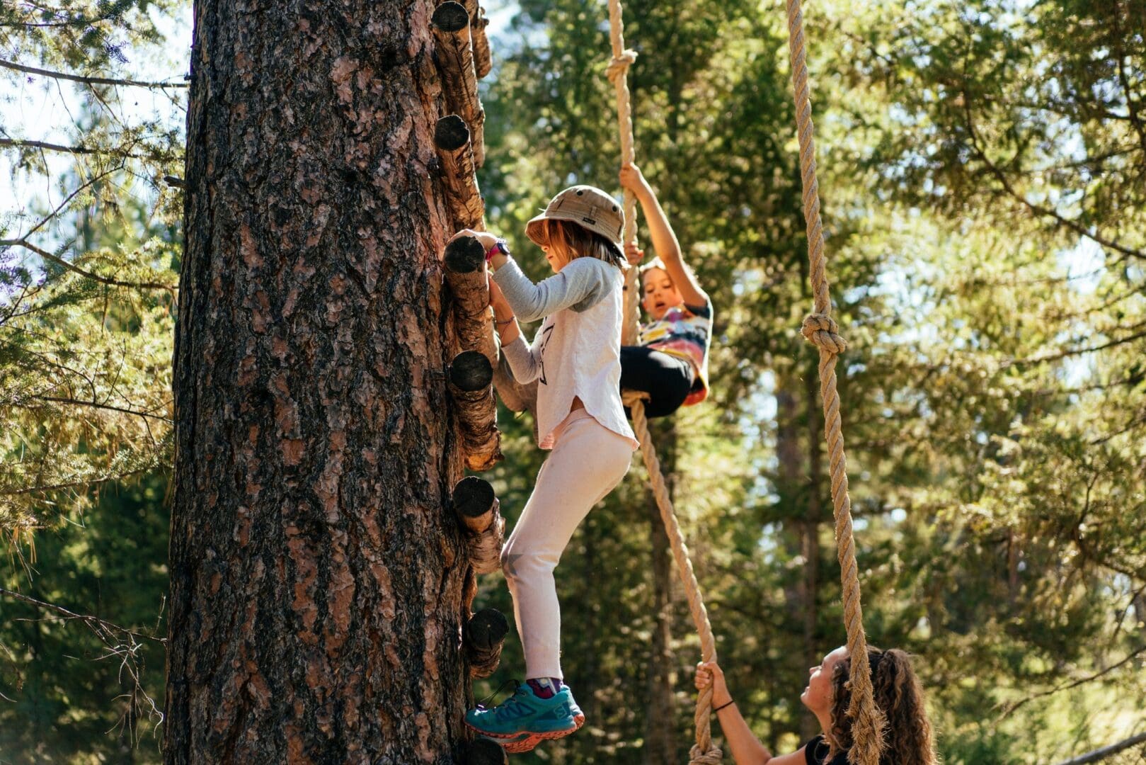 2 girls climbing up a forest tree with a rope