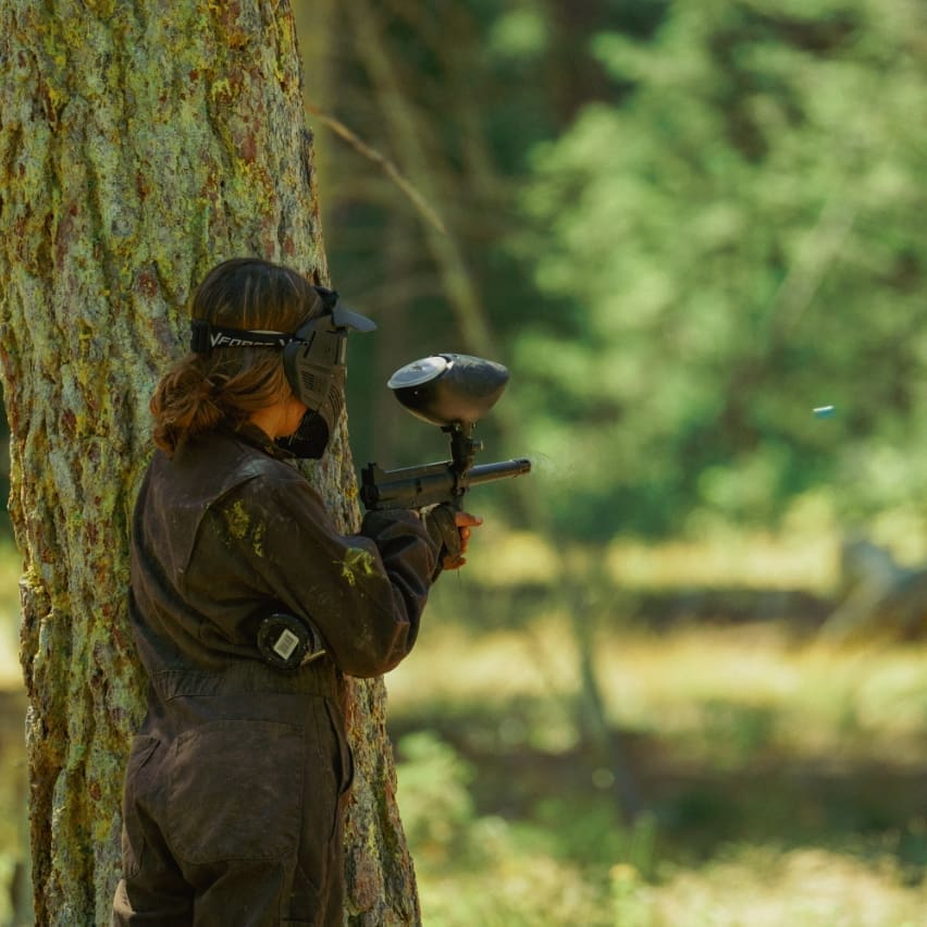 Woman shooting a paintball gun behind a forest tree