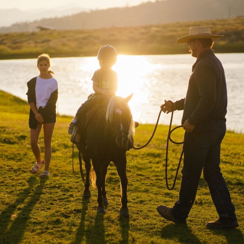 Child riding a pony beside a river being led by a wrangler